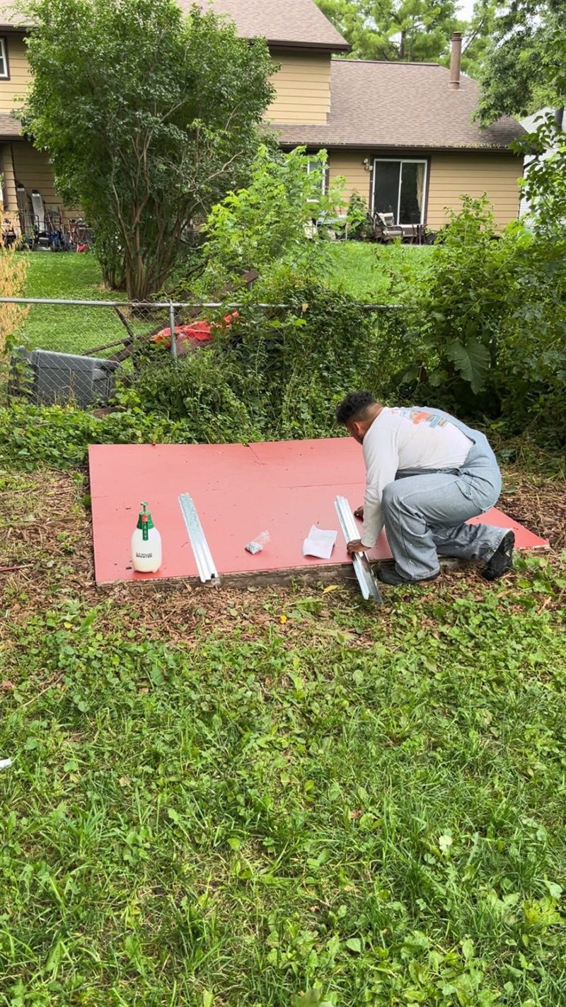 A worker laying out the base rails of a shed on a prepared platform in a back yard.