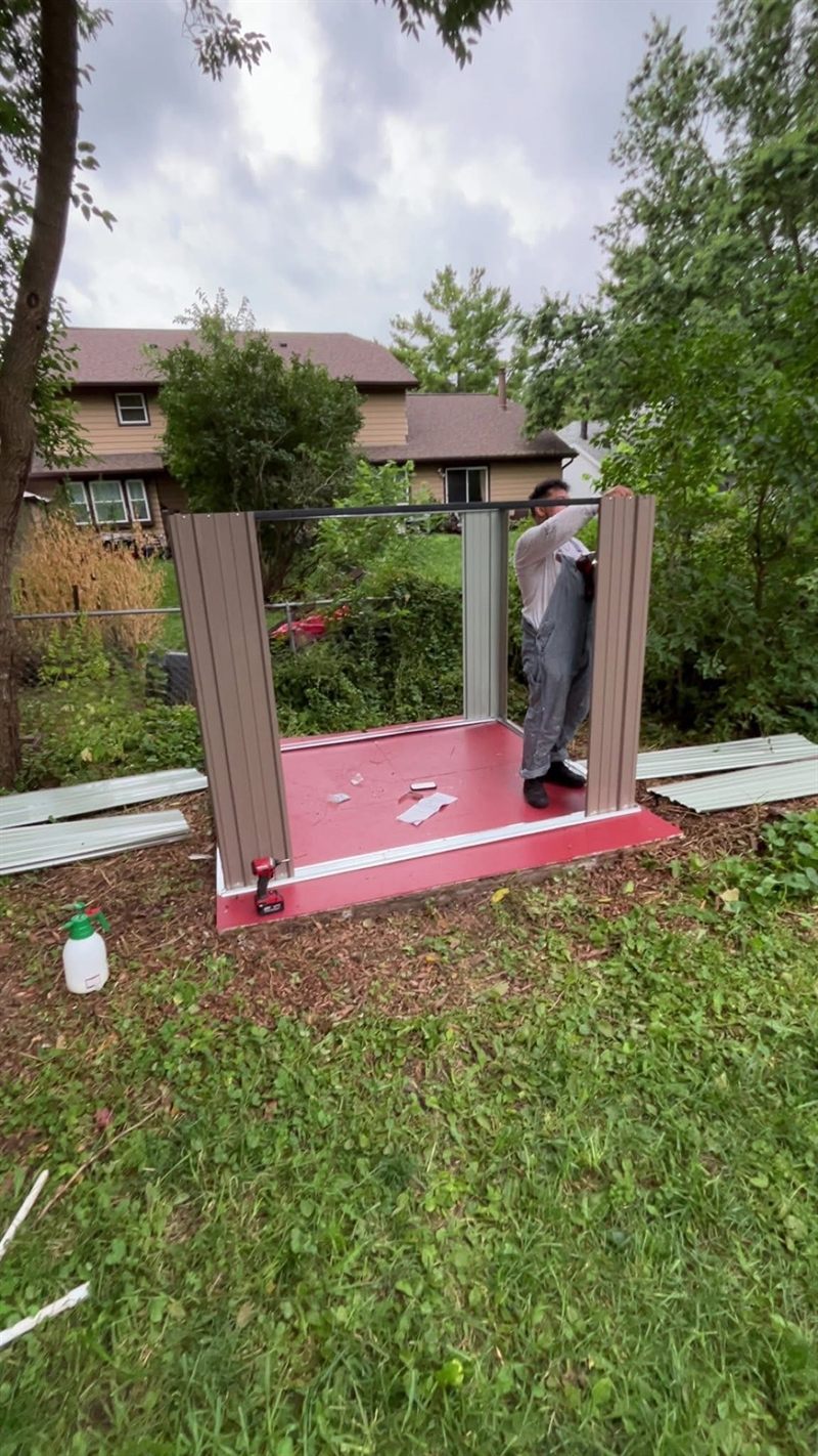 A metal storage shed being assembled on a prepared base in a back yard.